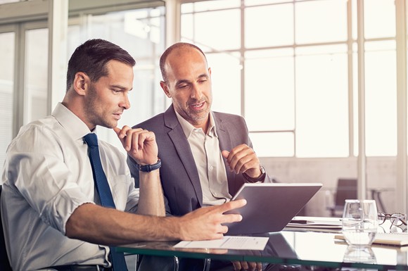 Two men in business-casual clothing, sitting at a table looking at a tablet