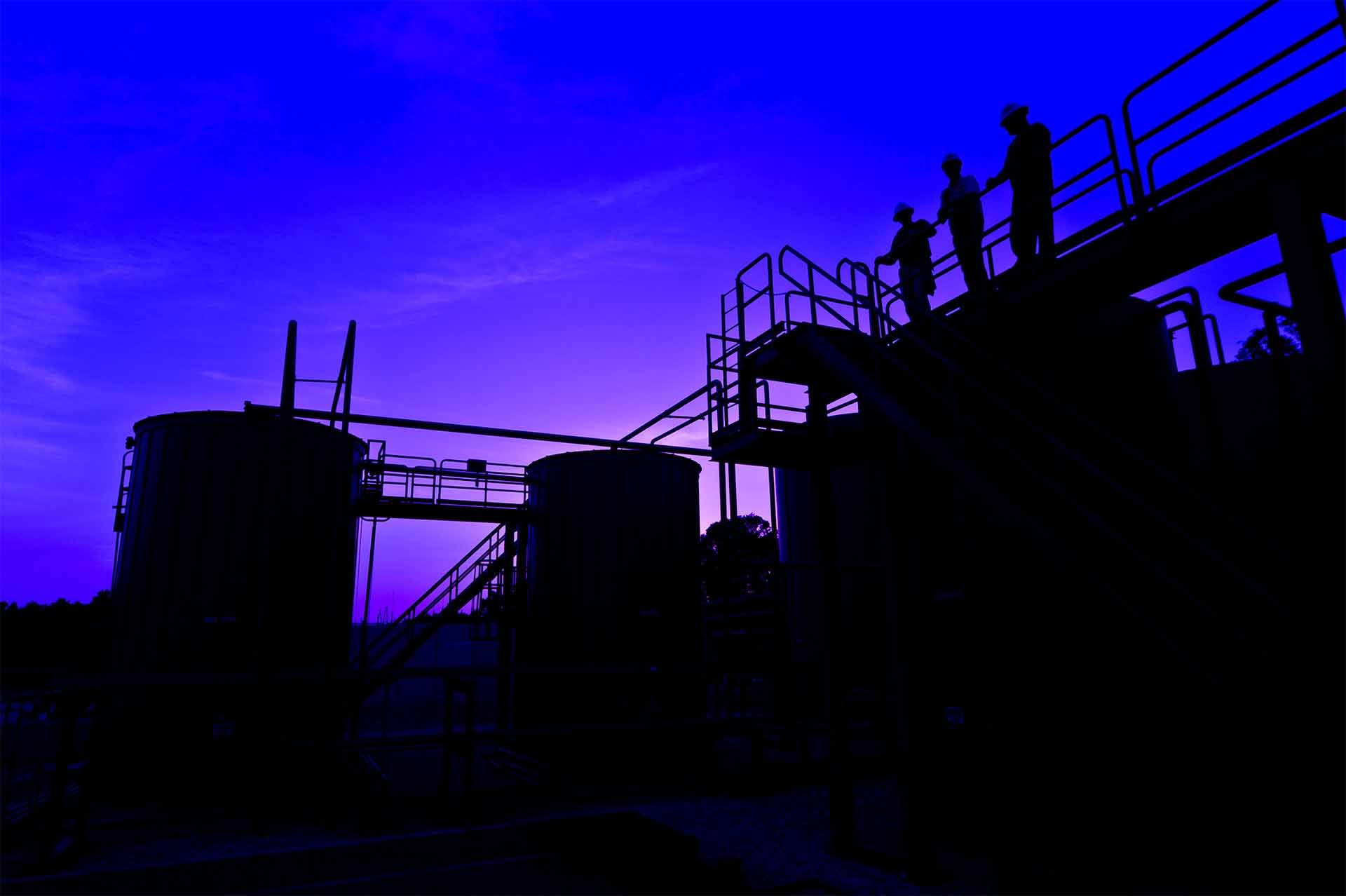 Three backlit workers at dusk on a railed walkway over an energy storage and pipeline facility.