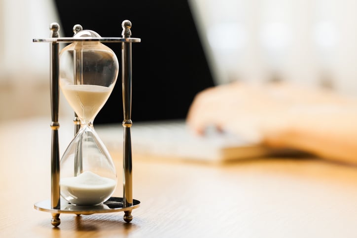 An hourglass sitting on a desk, with a laptop in the background