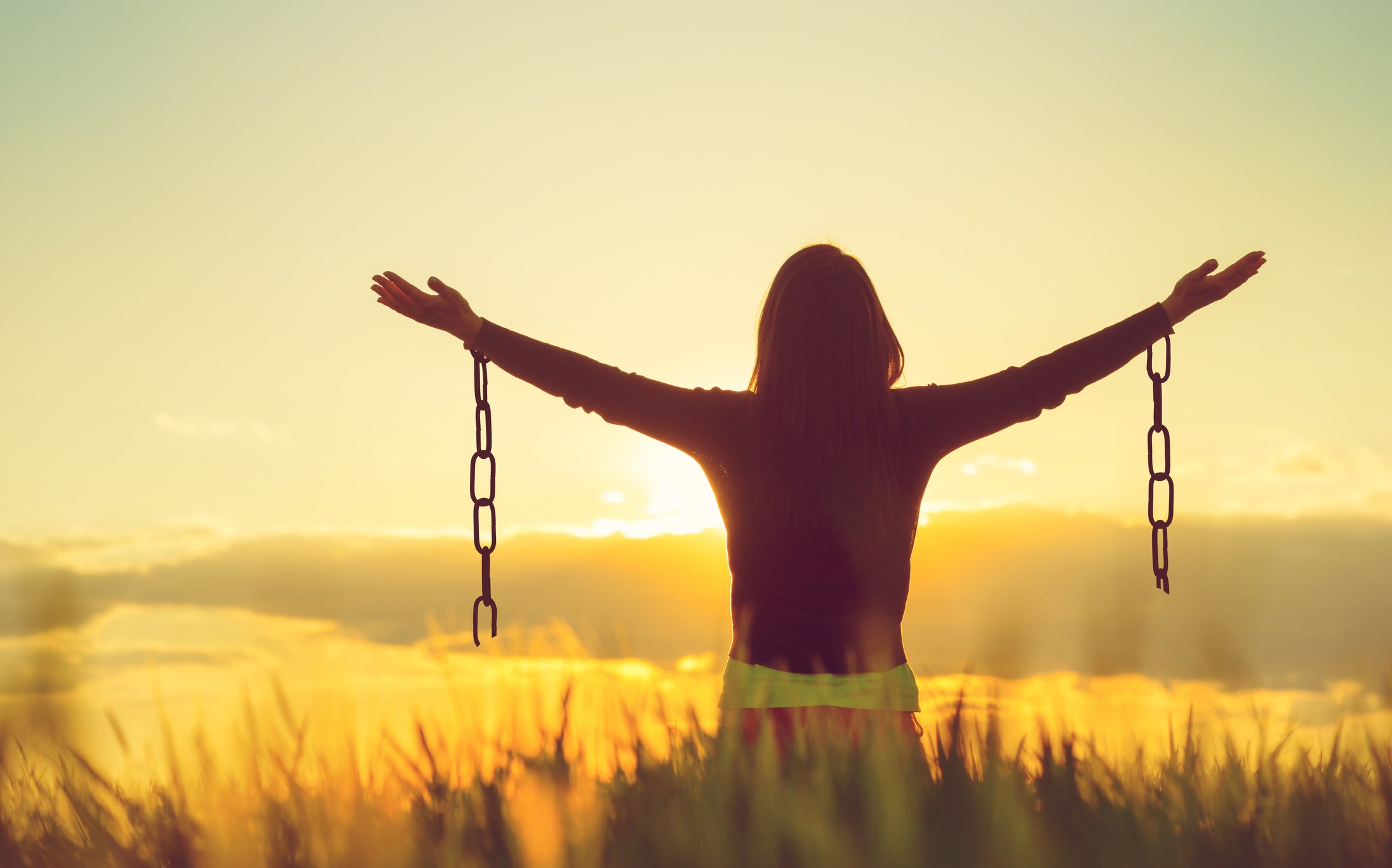 Woman in a field with arms raised, facing the sun, with shackles coming down from her wrists