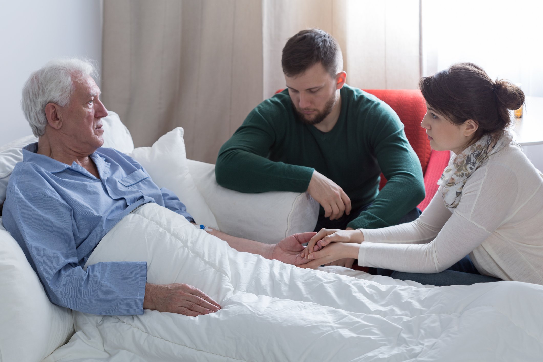 A sickly man in bed surrounded by his adult woman and man.