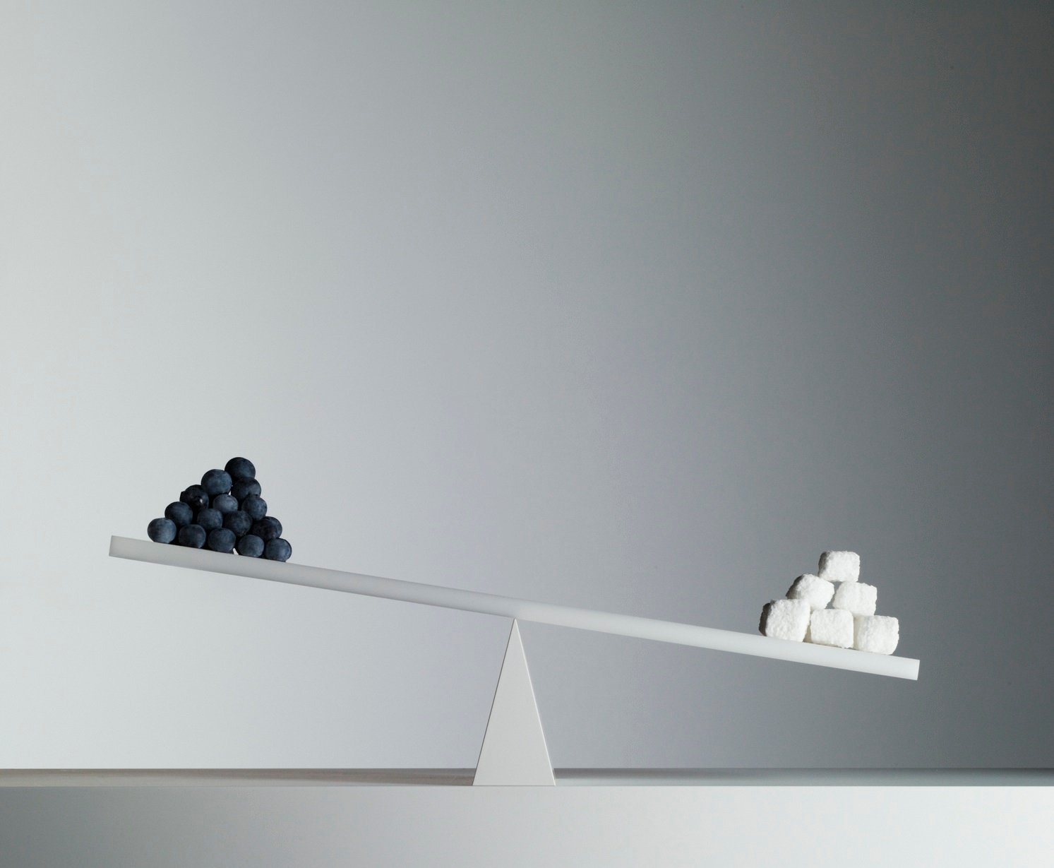 A seesaw with a pyramid of small black spheres on the left and larger white cubes on the right, against a blank gray wall.