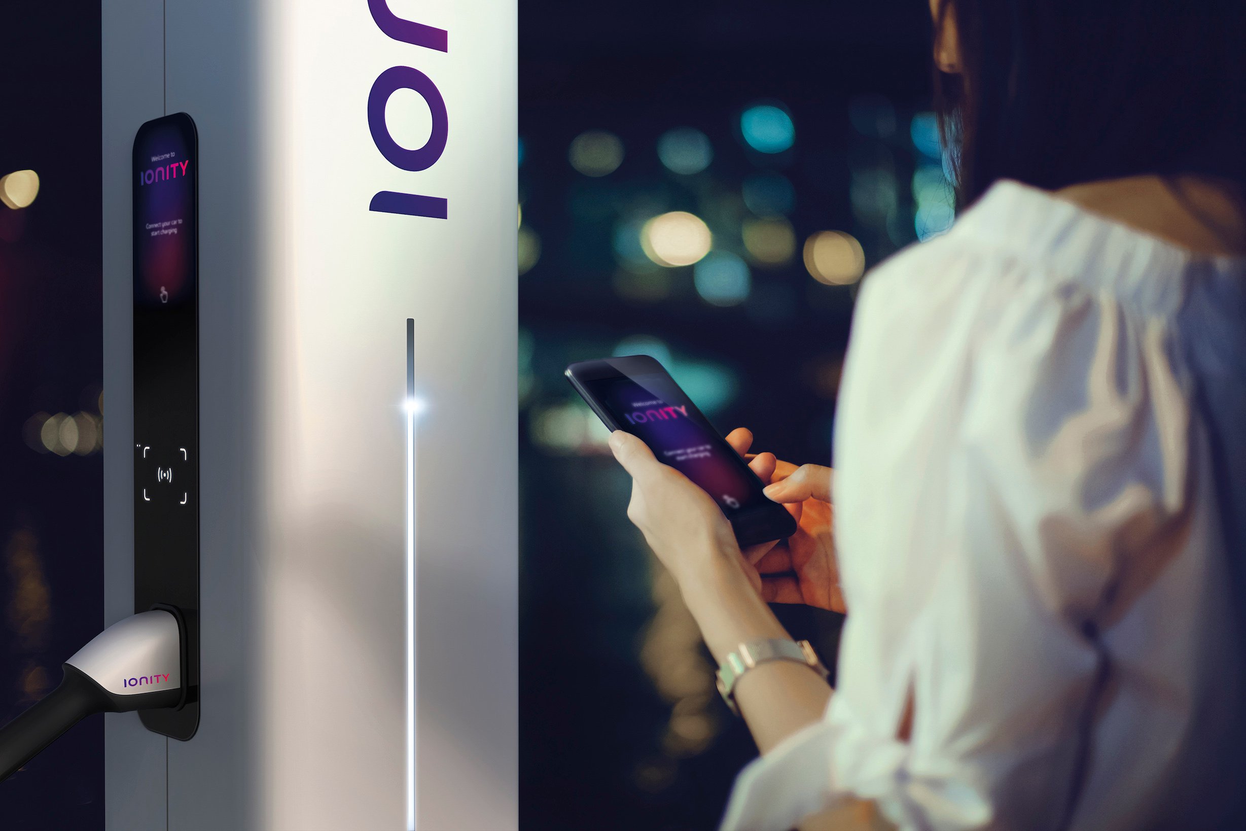 A woman is standing next to an Ionity charger, holding a cellphone displaying the company's app.