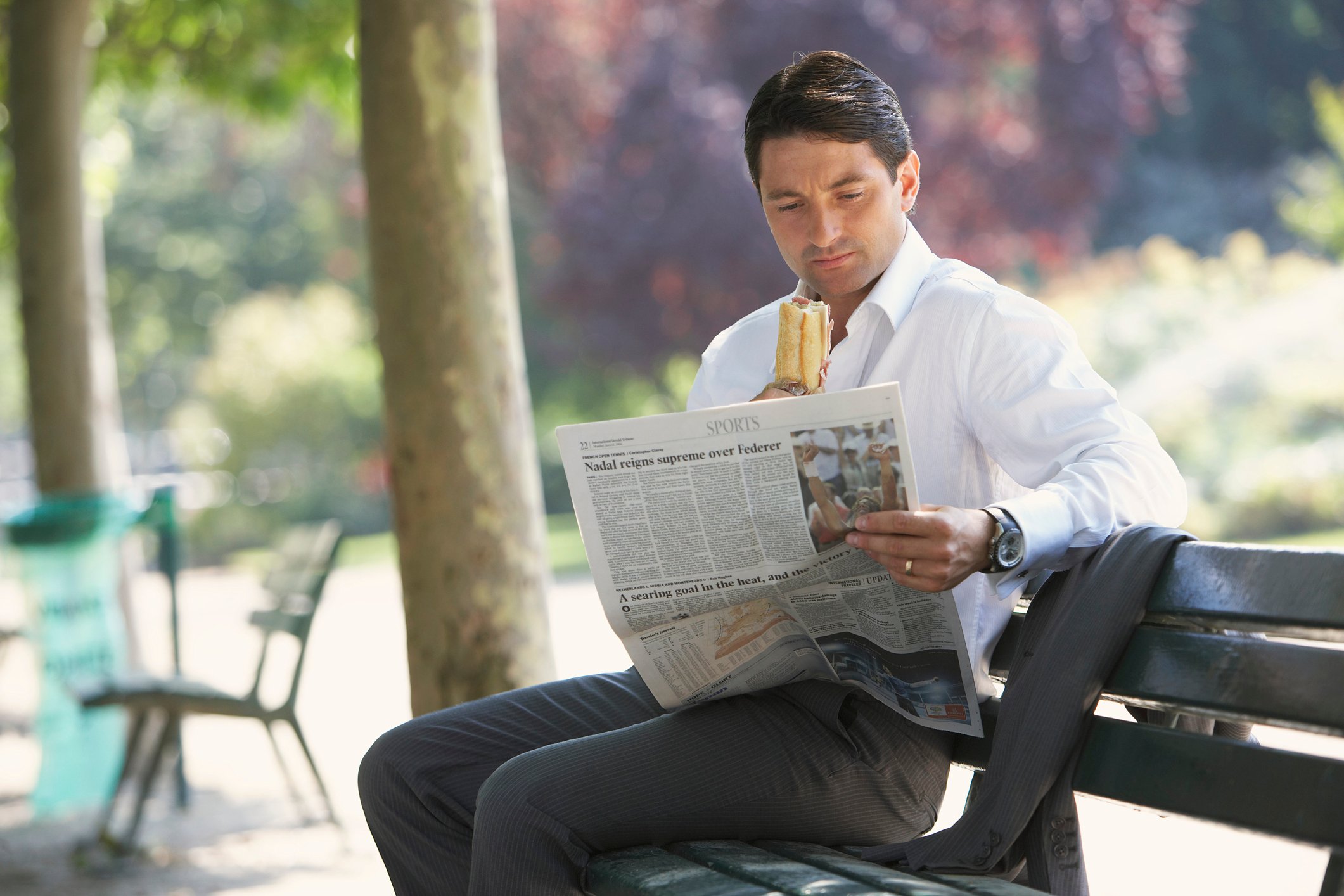 Man in professional clothing reading a newspaper on an outdoor bench while holding a sandwich.
