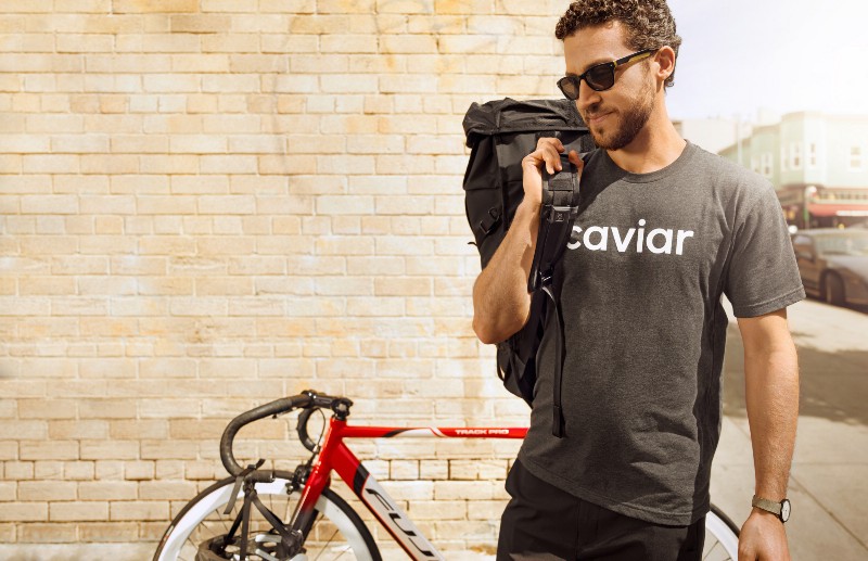 A Caviar employee carrying food in a bag over his right shoulder with a bike leaning on the wall behind him. 