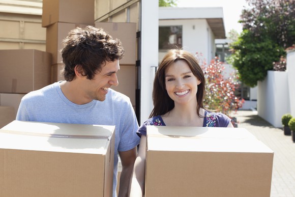 A young adult male and female carrying boxes from a truck parked in front of a home.
