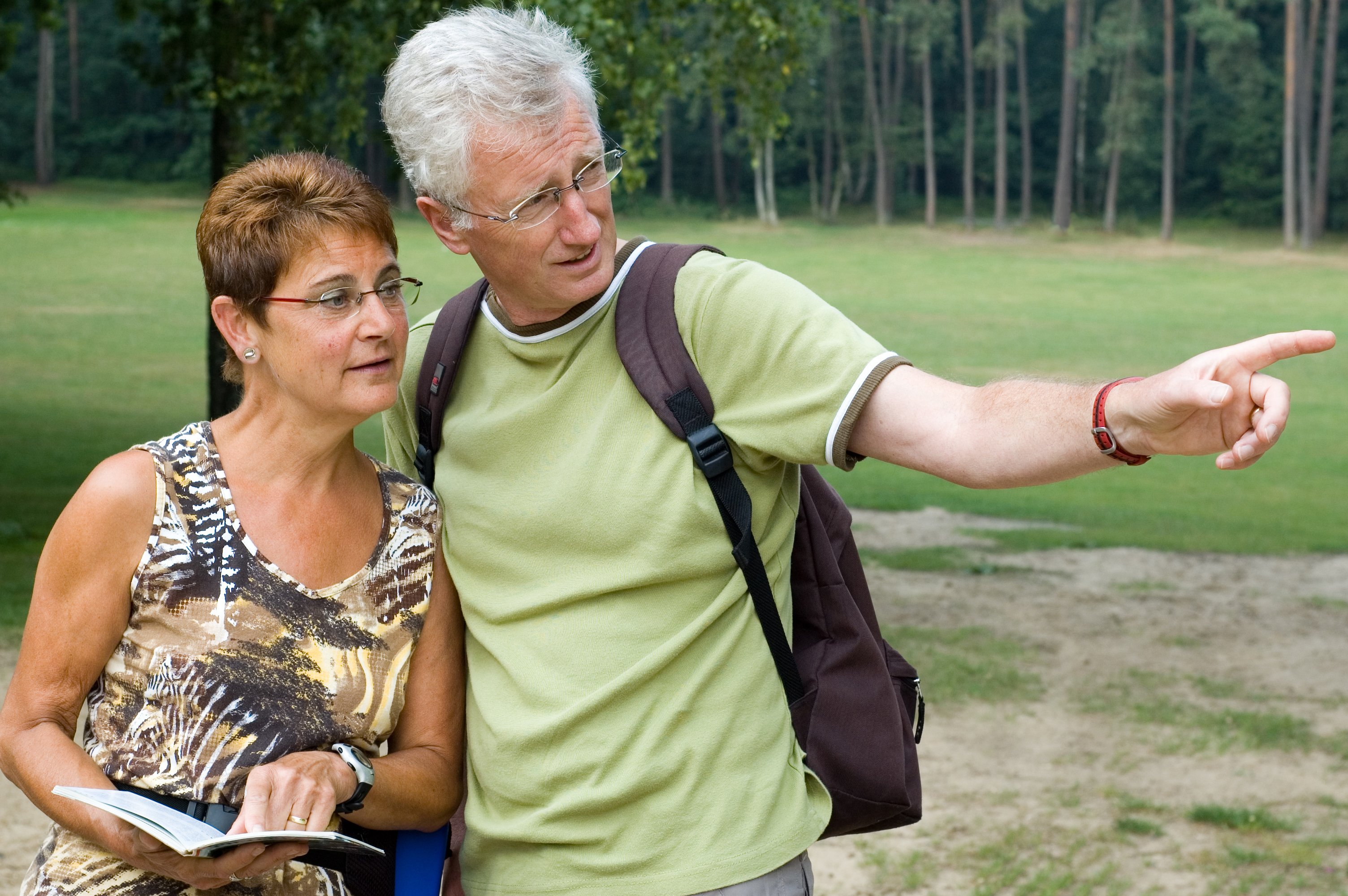 Senior couple outdoors, with woman holding a book and man pointing outward