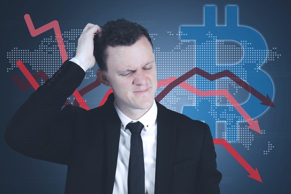 Man scratching his head in front of a large bitcoin logo and several falling arrows.