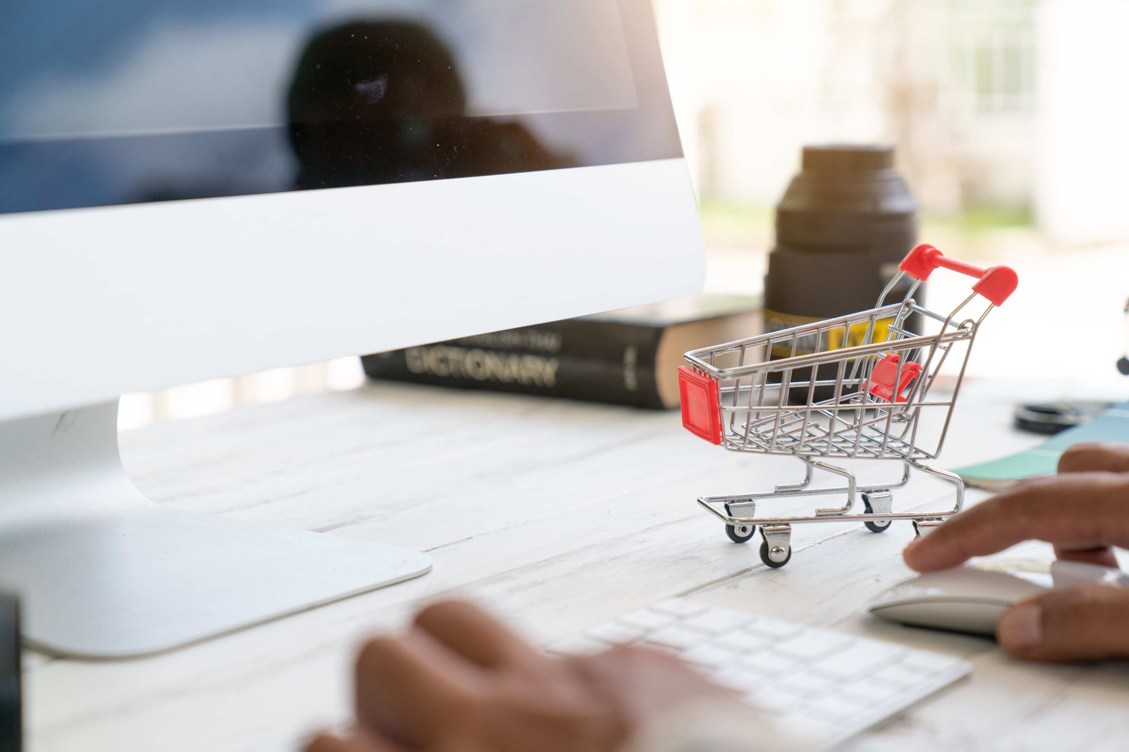 A tiny grocery cart sits on a desk next to a computer screen and someone using the keyboard and mouse.
