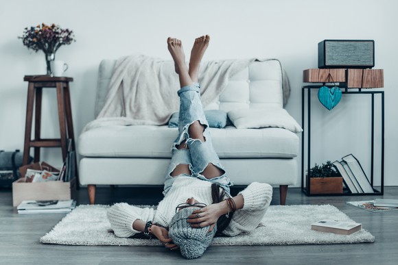A young person in casual fashion wear touching their head with their hand while lying on the floor with their feet up on a couch.