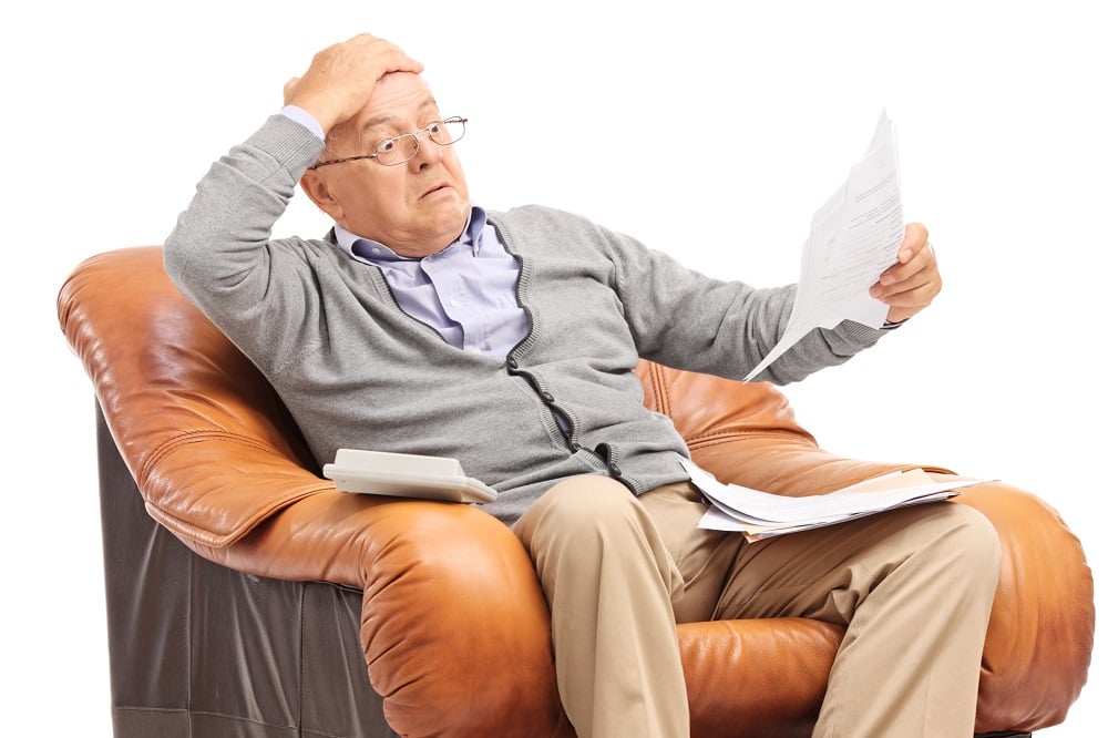 Older man sitting in a leather armchair looks at a paper with a shocked look on his face.