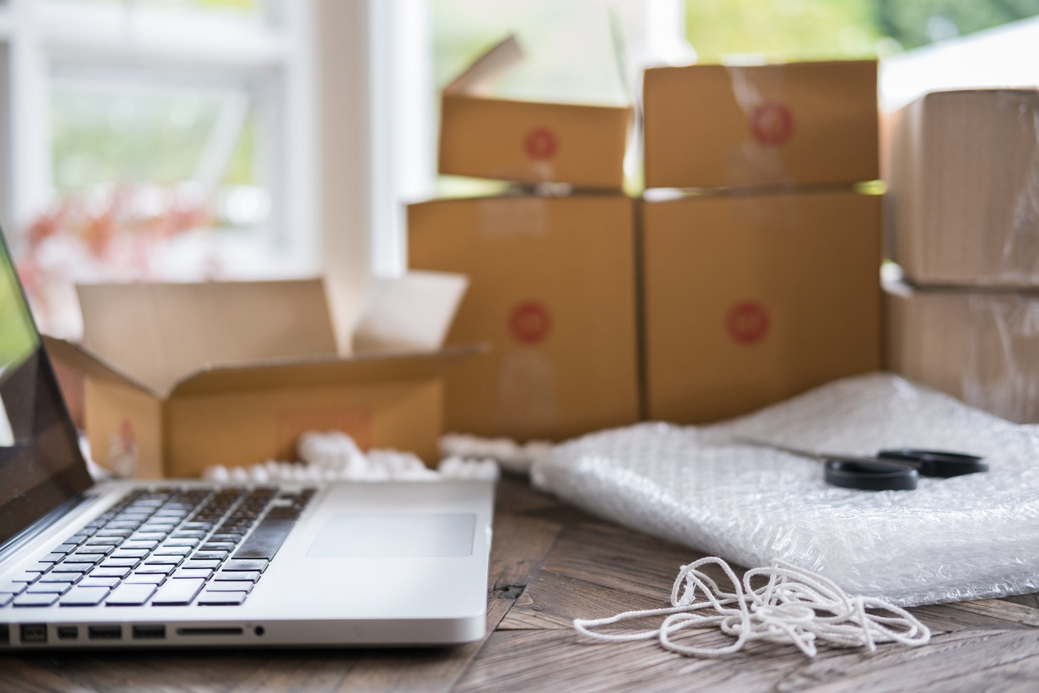 An open laptop on a table with boxes, bubble wrap, scissors, and string.