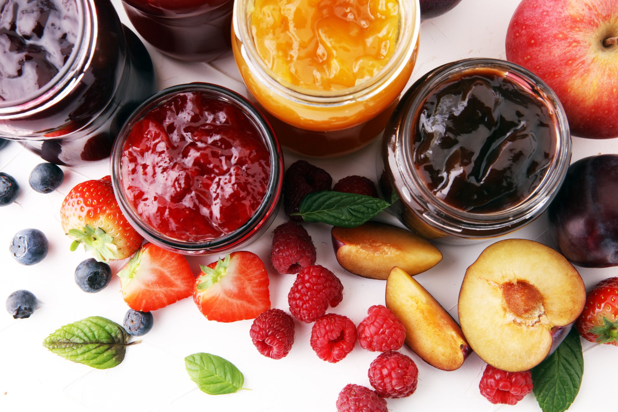 Four jars of different types of jelly viewed from above next to strawberries, raspberries, blueberries, mint leaves, an apple, and a peach