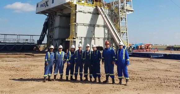 Eight workers wearing hardhats in front of a drilling rig on sandy ground under a clear blue sky.