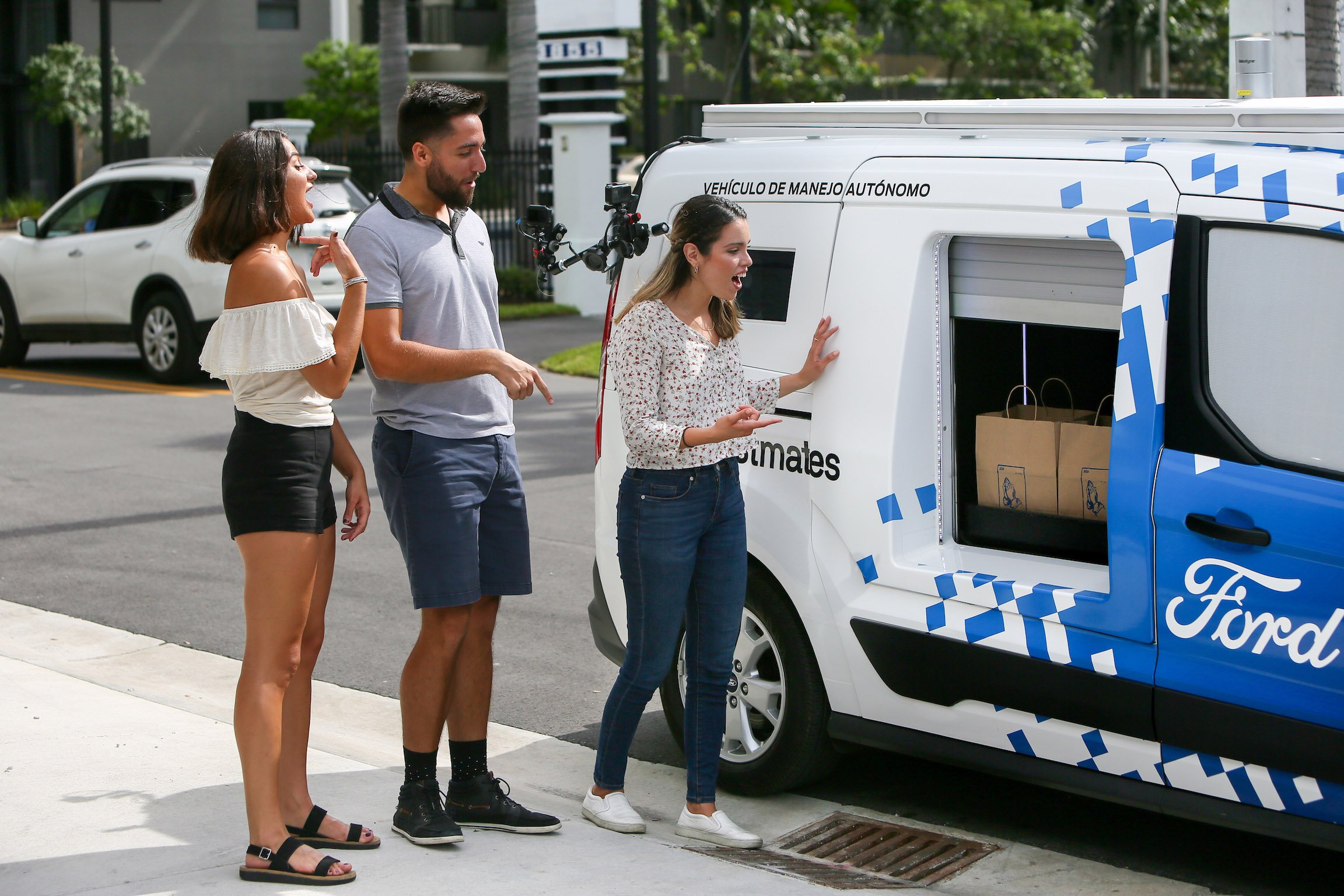 The modified Ford Transit Connect is shown parked at a curb with one of its lockers open, and three people are waiting to take the contents.