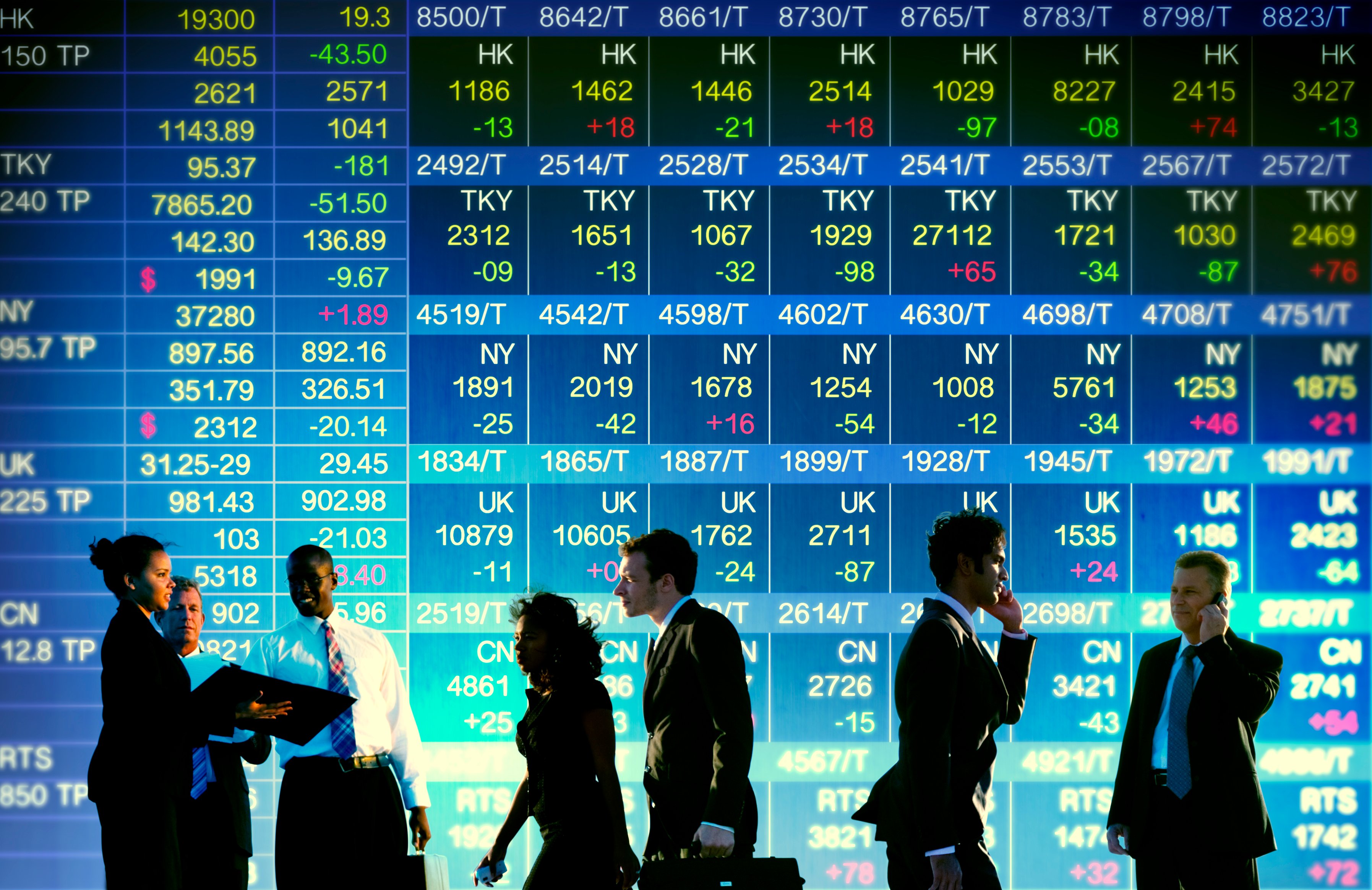 Men and women in suits, standing and walking in front of a display of stock prices.