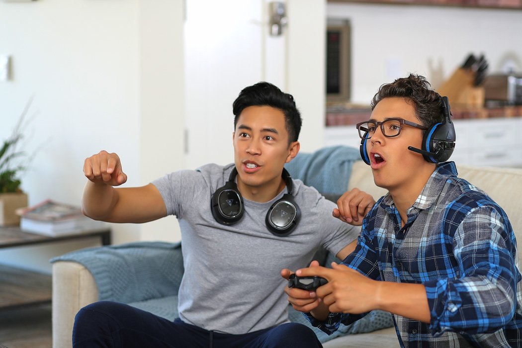 Two young men sitting on a sofa -- one playing a console video game and wearing Turtle Beach headsets and the other cheering him on. 
