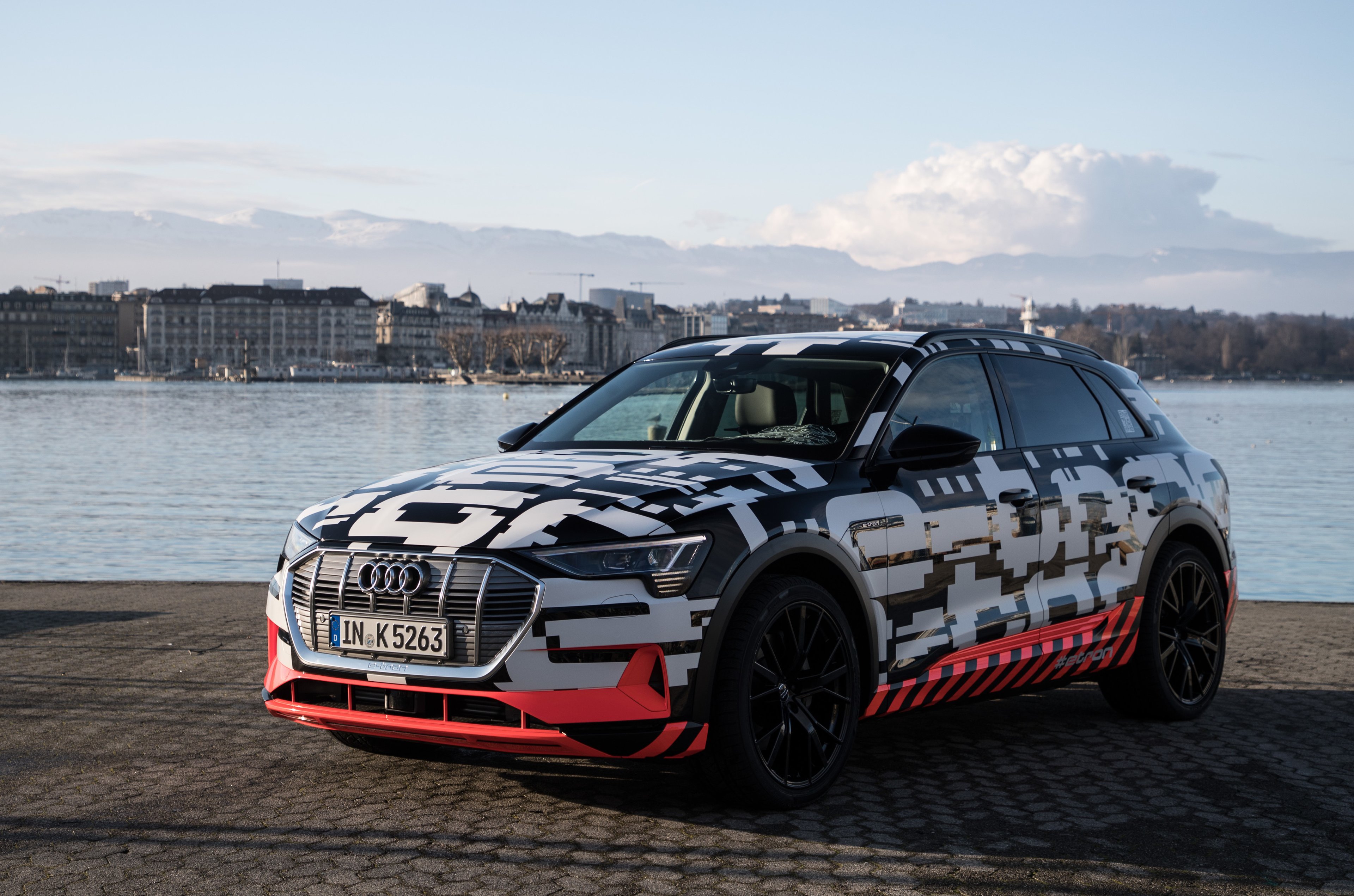 The e-tron quattro prototype, a sleek midsize SUV covered in black and white camouflage, parked on a lakefront. 