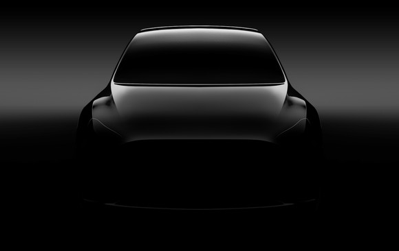A "teaser" photo of a silver Tesla Model Y, a sleek SUV, viewed from the front.