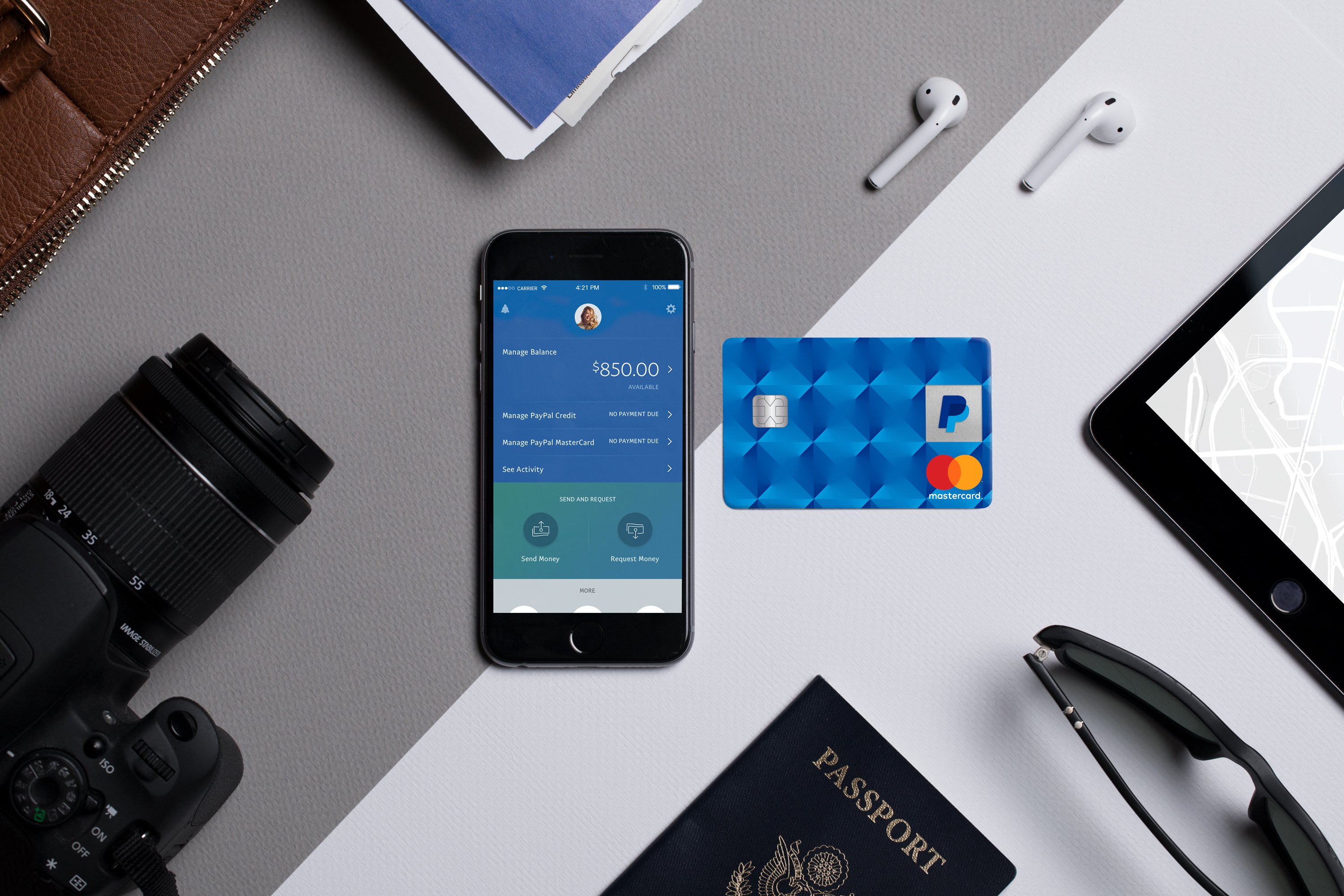 The PayPal-branded credit card on a desk next to a phone with the PayPal app, a passport, camera, earbuds, and sunglasses.