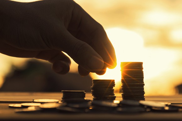 A hand placing a coin on a stack of coins with the sun in the background.