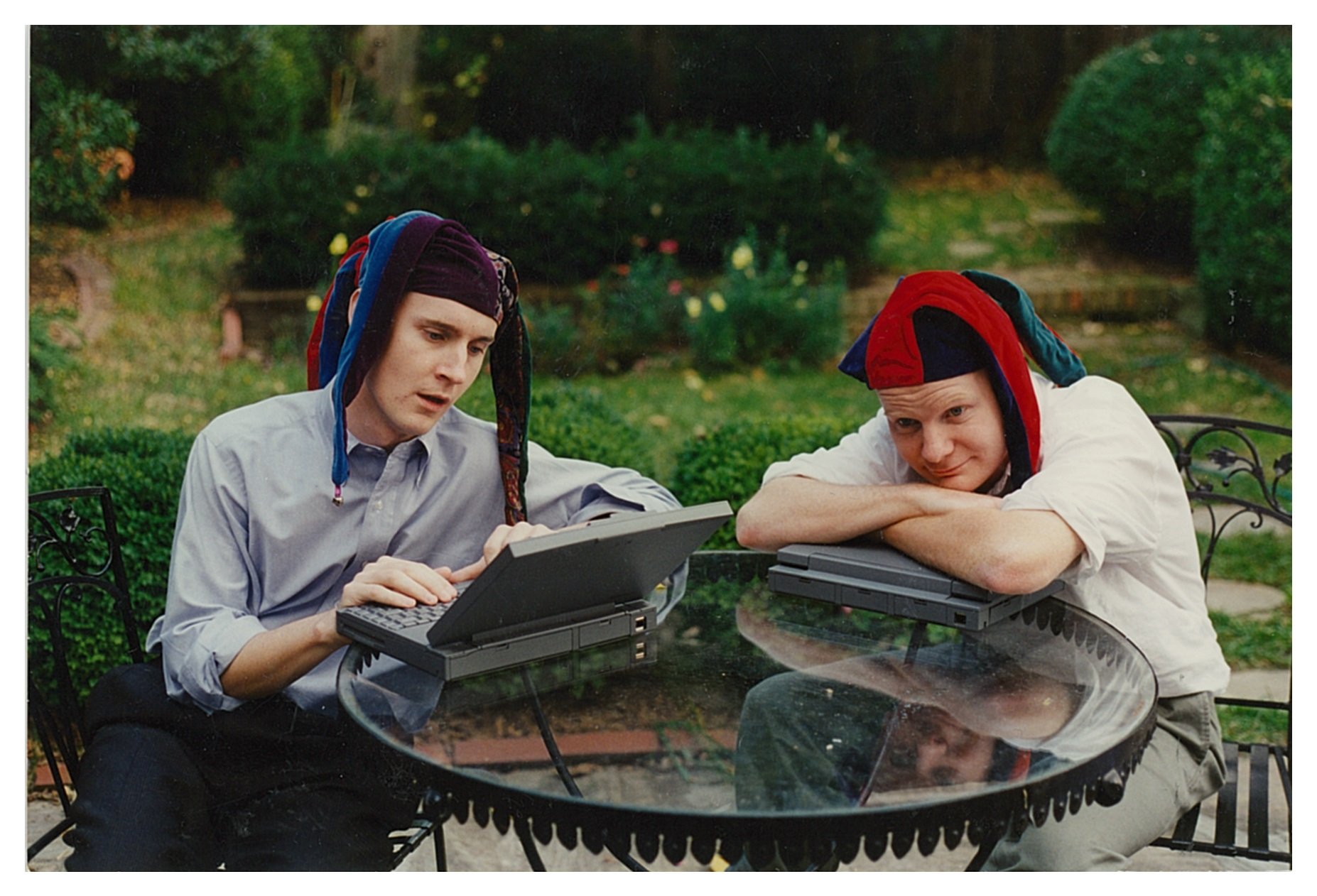 Two men in jester caps at a table. One is typing on an old laptop, wearing a jester cap and a blue shirt. The other has his arms folded over his computer and is resting his face on them, wearing a white t-shirt and a jester cap. Bushes in the background.