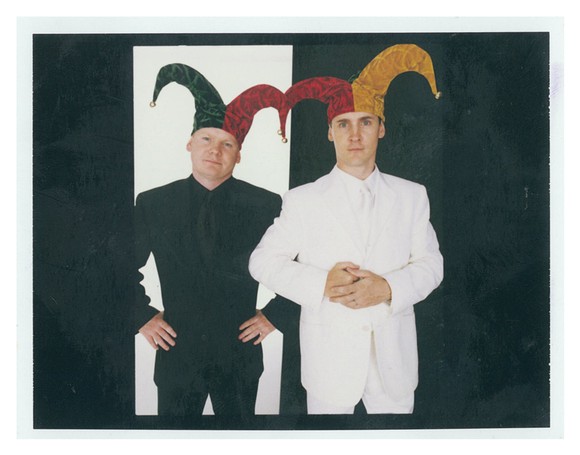 Two men (Tom and David Gardner), one in an all-black suit and the other in an all-white suit, both wearing jester caps, in front of black and white backgrounds.
