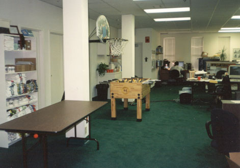 Room in an office building -- bookshelf with towels and books stacked on it, a basketball hoop, an empty brown table, and a foosball table. Desks in the corner.