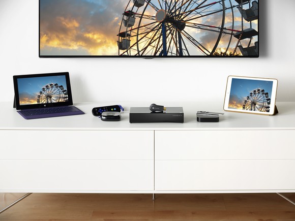 Various computing devices on a table with a television mounted on the wall.