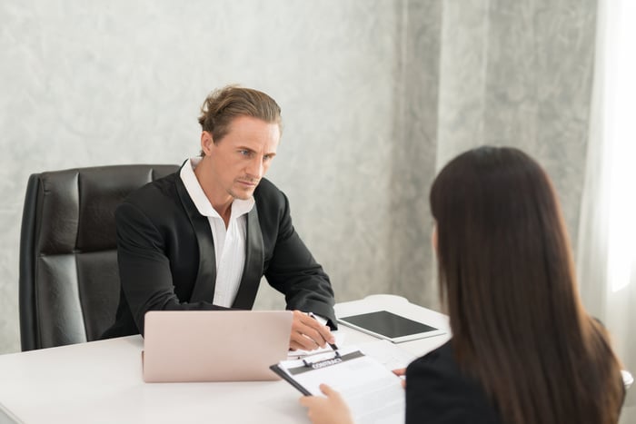 Professional male with stern expression sitting across from a professional woman holding a clipboard