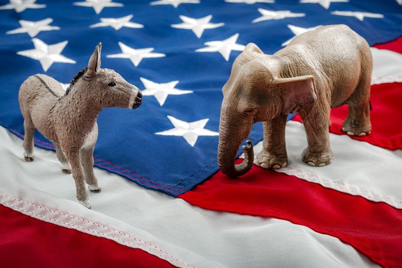 A Democrat donkey and a Republican elephant having a stare-down while standing on the American flag.