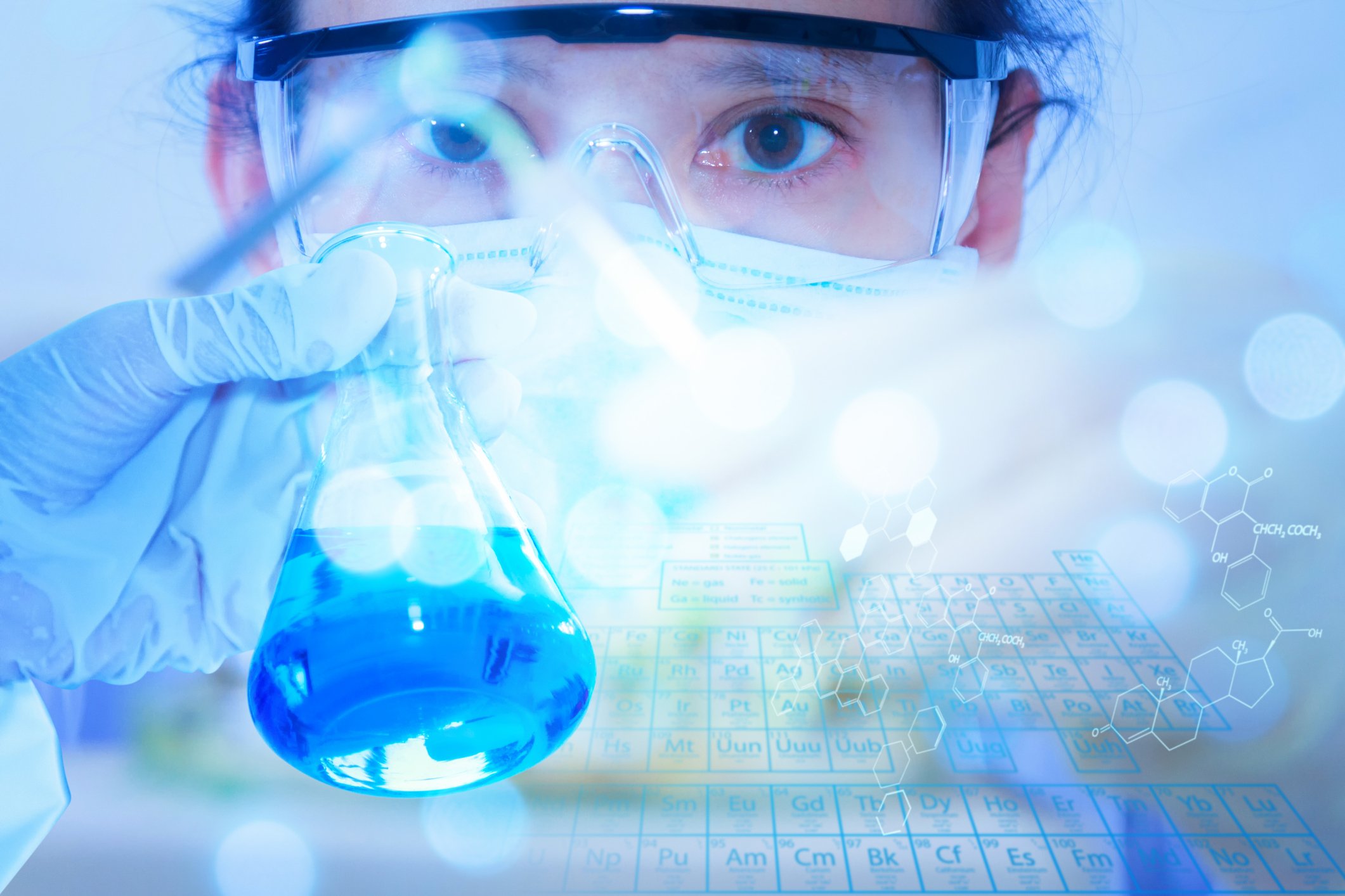 Gloved hand holding a beaker in front of the face of a scientist wearing safety eyeglasses