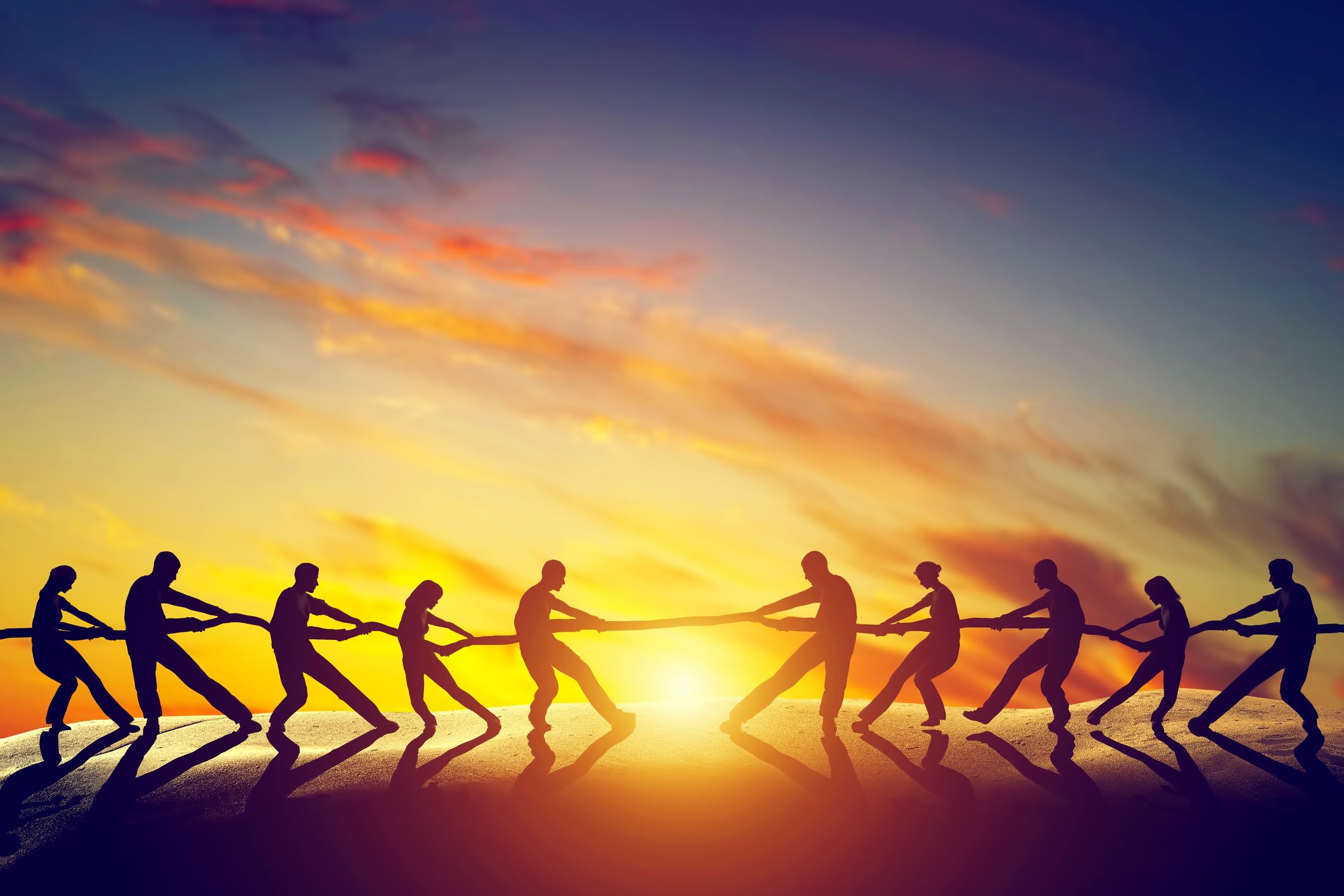 Two groups of people pulling a rope, playing tug of war, with the sunset as a backdrop