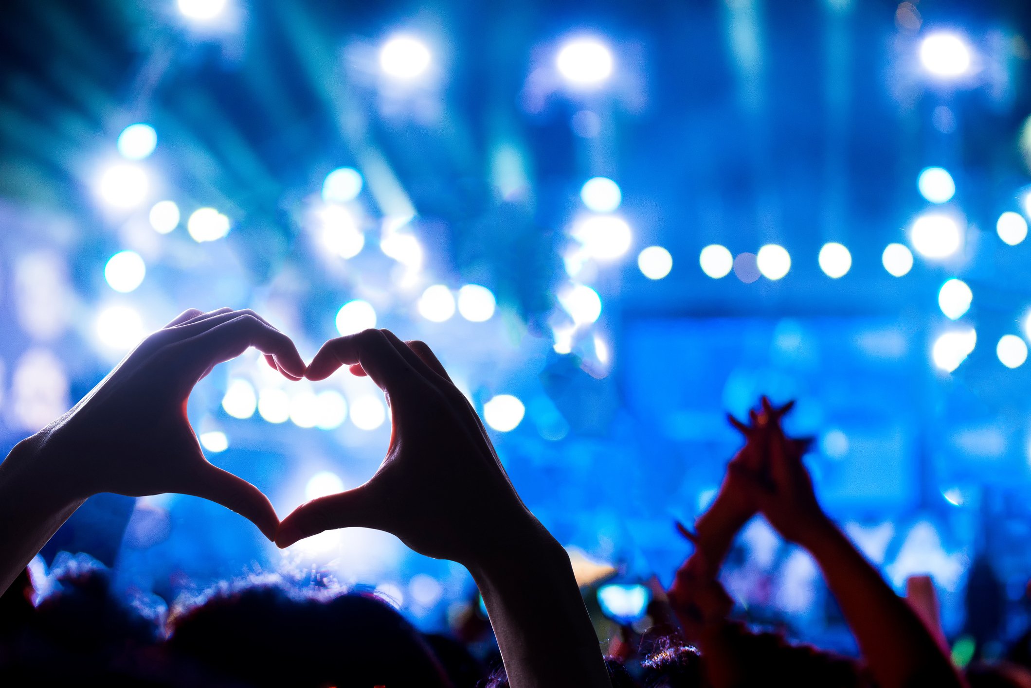 Crowd at a rock concert, where the view focuses on one audience member forming a heart sign with her hands.
