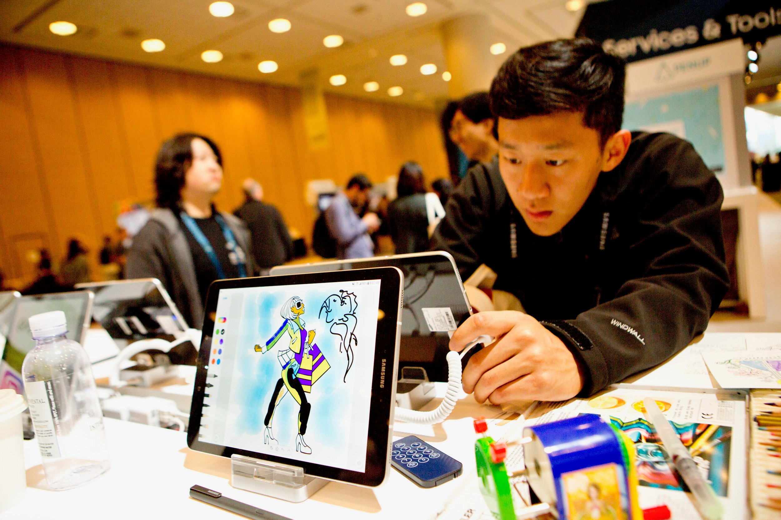 A Samsung tablet in the foreground with a person looking at a Samsung tablet in the background.