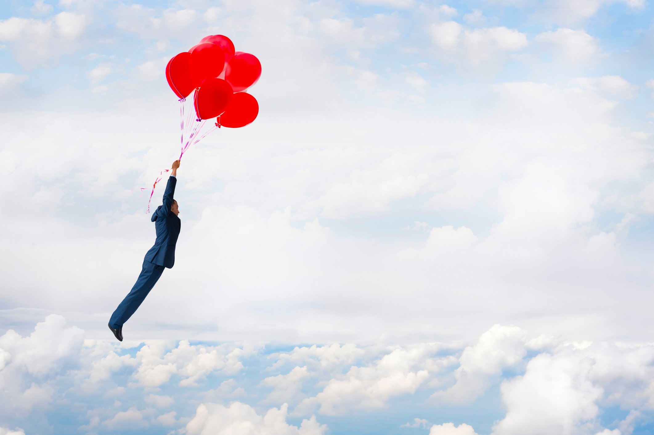 Man in a suit flying above the clouds by holding on to a cluster of red balloons.