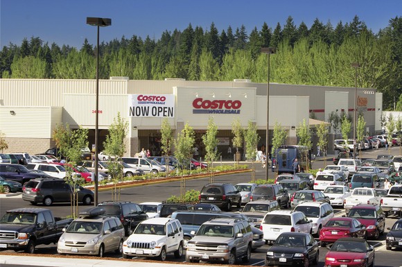 The front of a Costco warehouse with a full parking lot, and a sign reading "Now open."