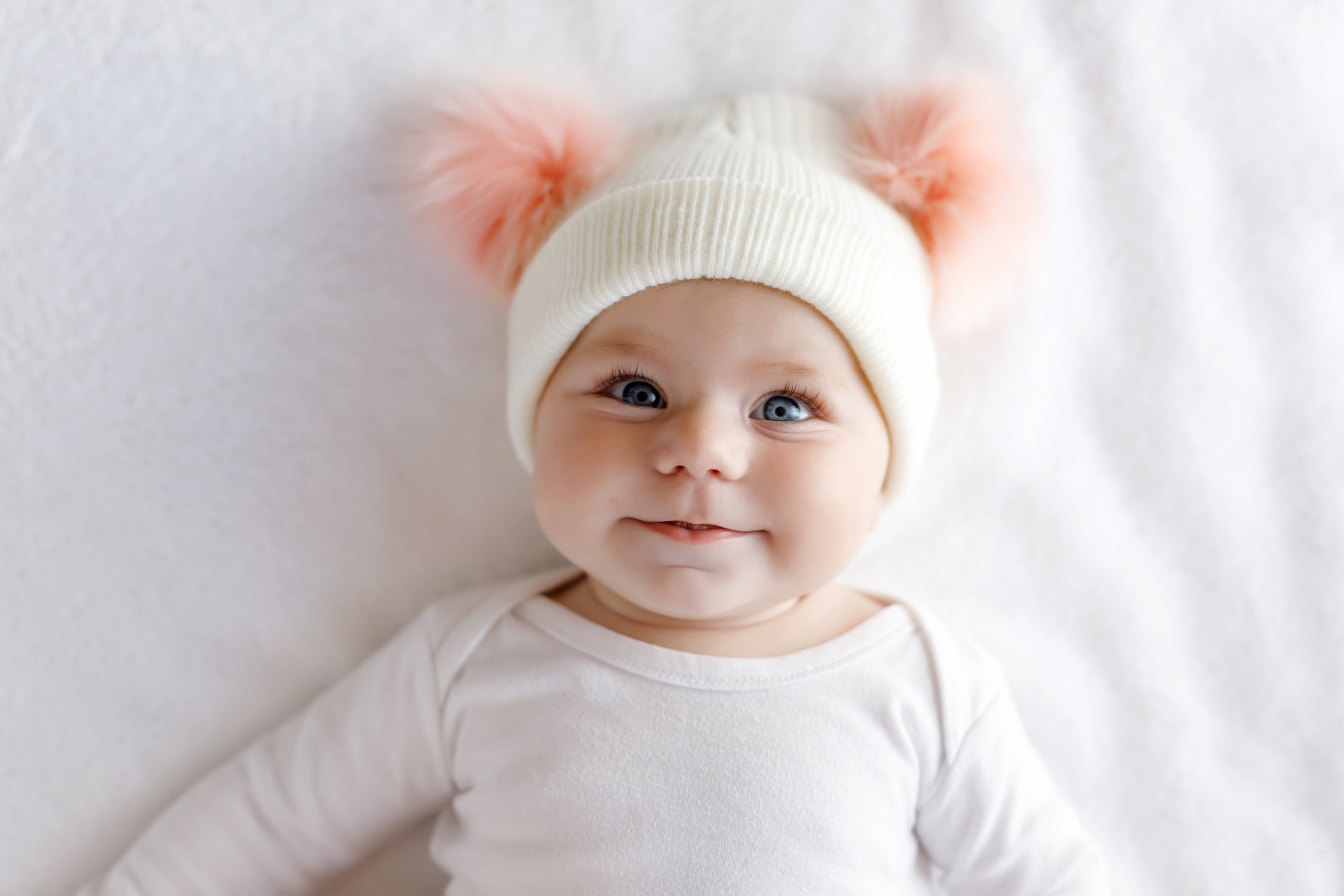 A baby wearing a white knit hat and white shirt while lying on a white sheet.