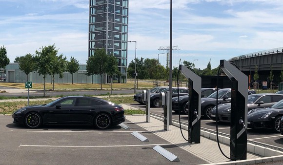 Four electric-vehicle chargers in the parking lot of a Porsche dealership. A black Porsche Panamera E-Hybrid is plugged into one of the conventional chargers. 