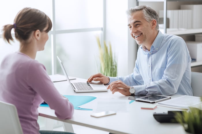 Smiling female sitting across the desk from a smiling older man who's typing on a laptop