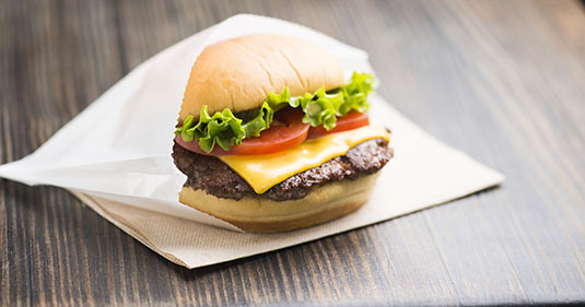 Shake Shack cheeseburger with tomato and lettuce tucked into a waxed paper bag, sitting on a napkin