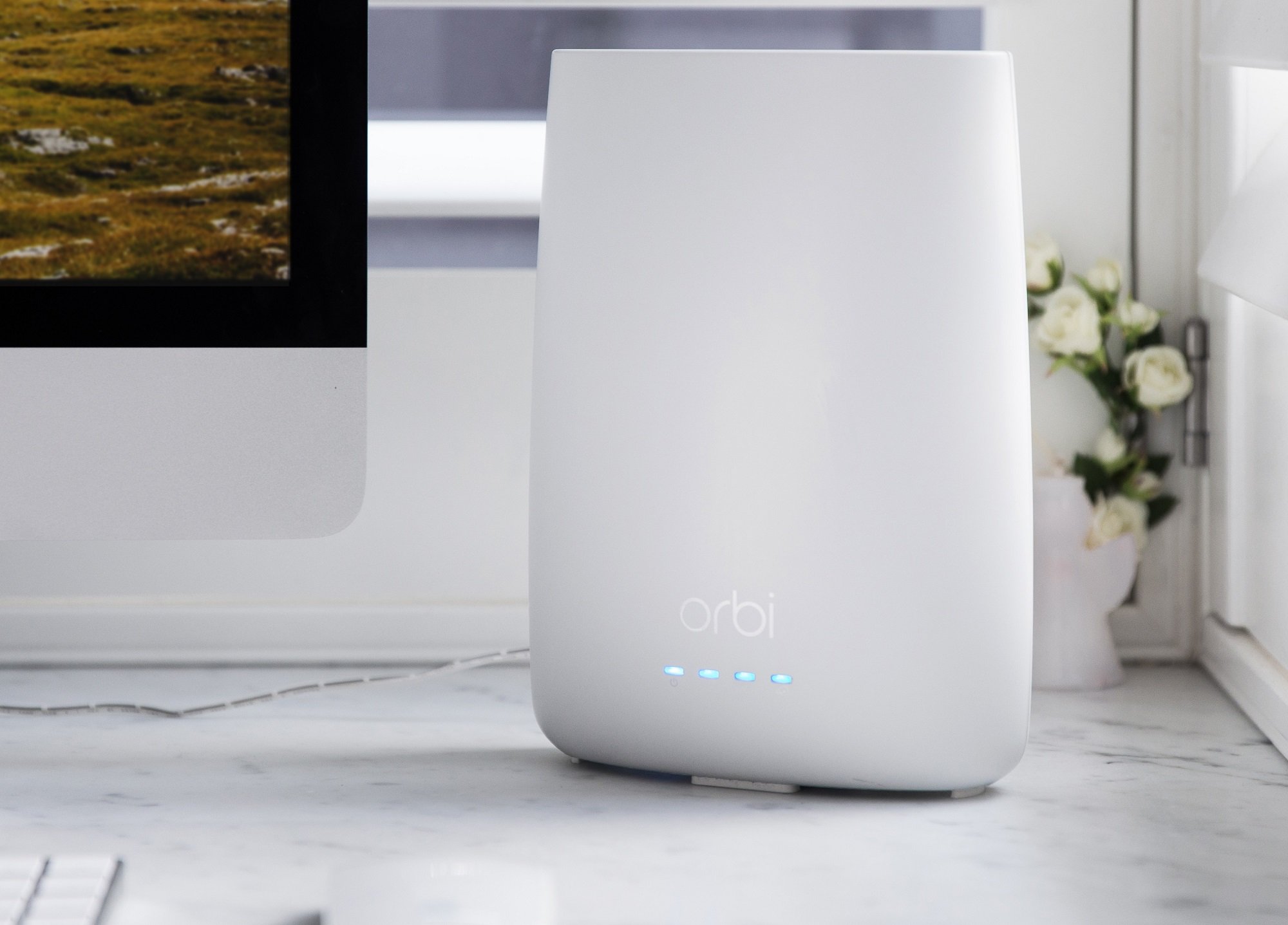 White Netgear Orbi cable modem and Wi-Fi router sitting on desk next to computer monitor