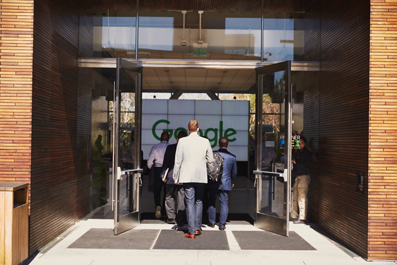Several well-dressed people walking into an open door with the Google logo prominently displayed.