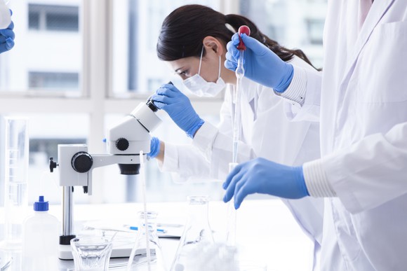 Female scientist looking through microscope with another scientist holding a dropper and test tube