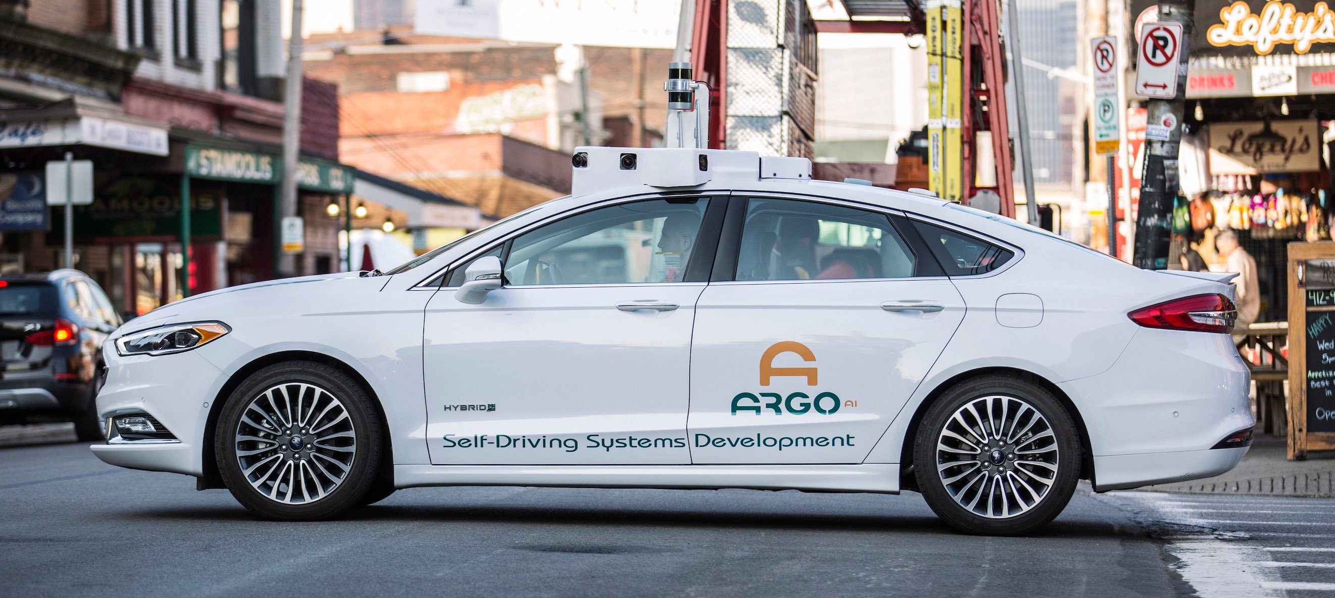 A white Ford Fusion sedan with "Argo AI" markings and visible self-driving hardware is shown on the streets in Pittsburgh.