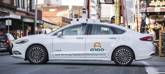 A white Ford Fusion sedan with "Argo AI" markings and visible self-driving hardware is shown on the streets in Pittsburgh.