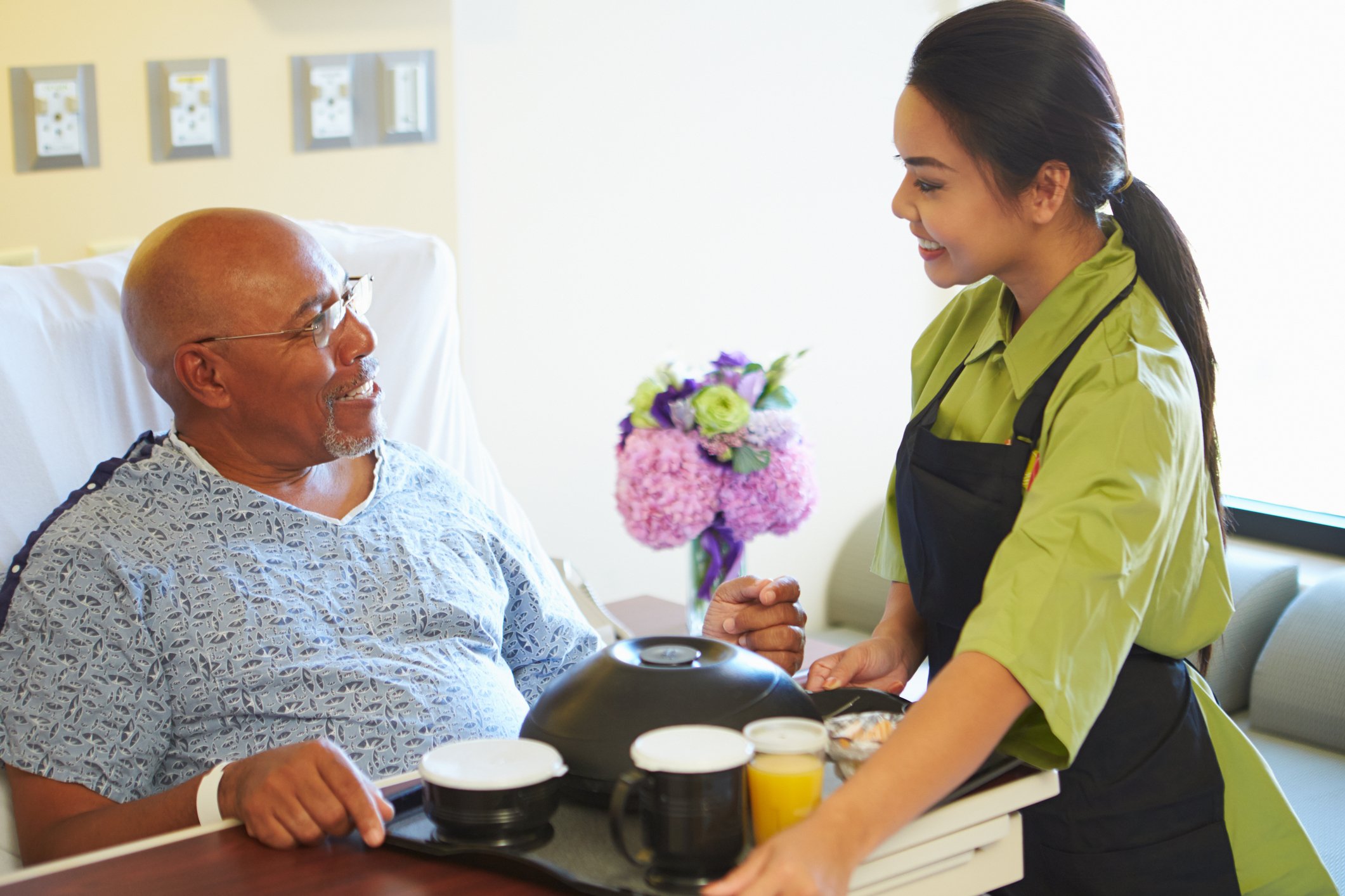Woman wearing an apron serves a tray of food to person in a hospital bed
