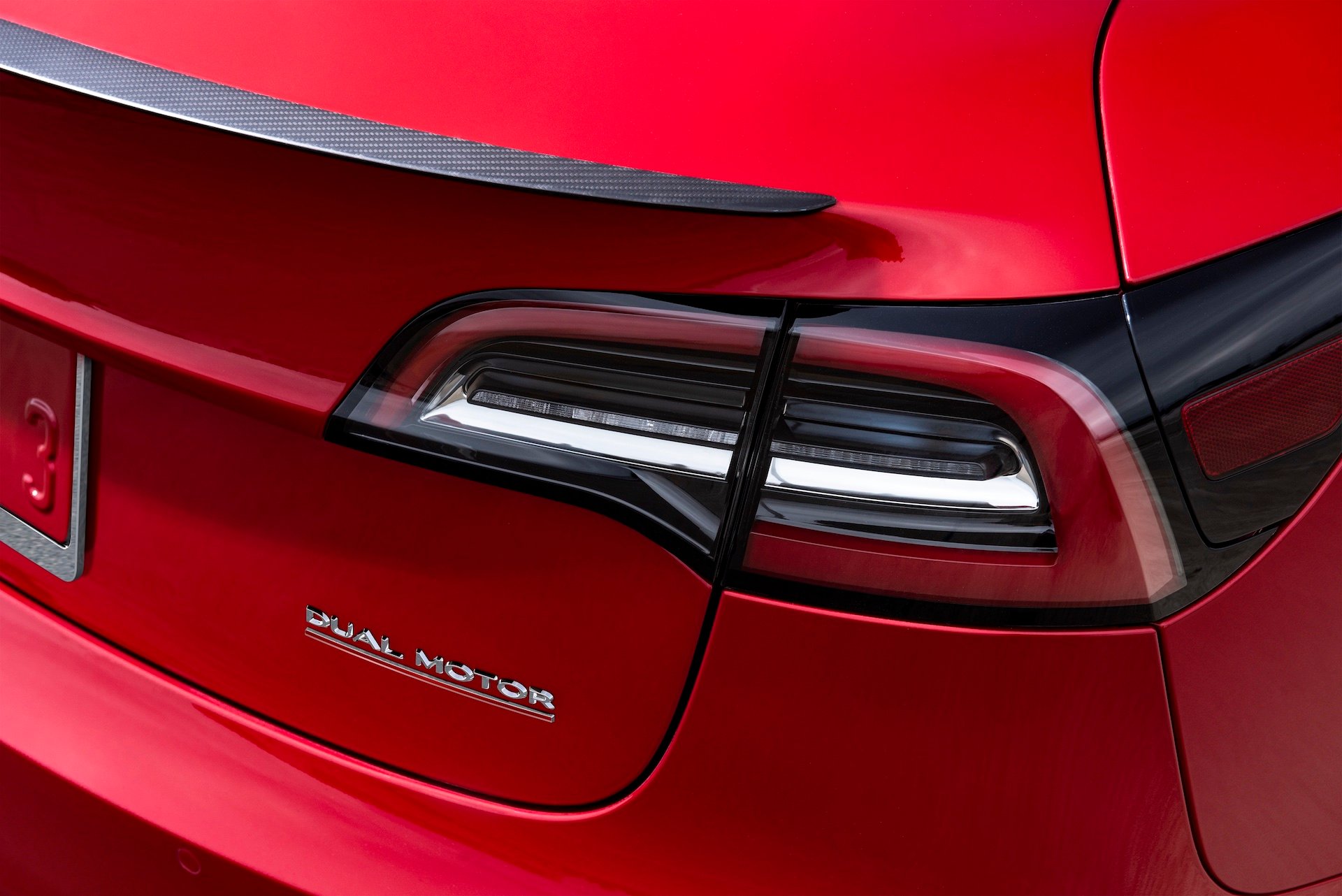 A close-up of the "Dual Motor" badge on the back of a red Tesla Model 3 sedan