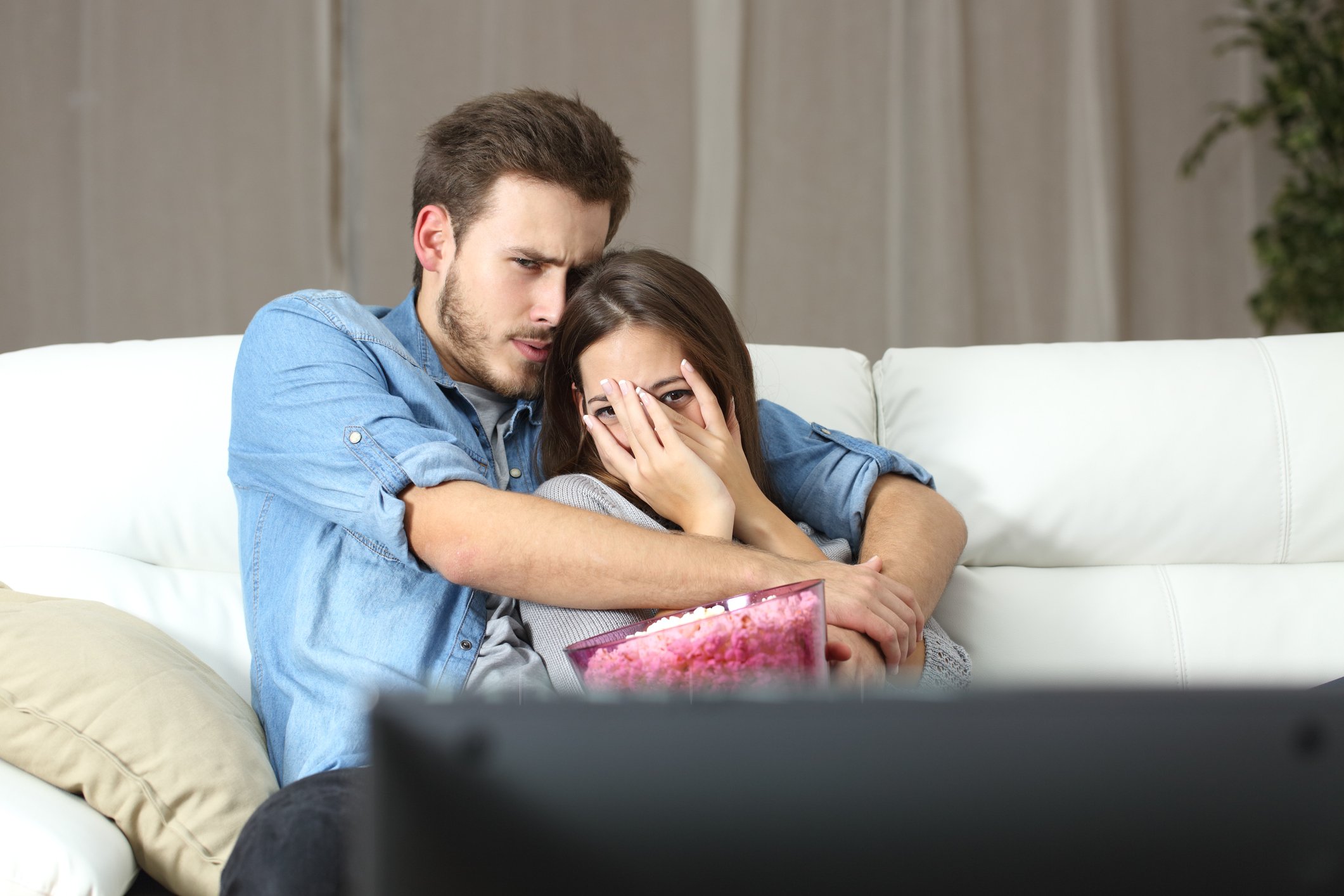 Young couple on a couch watching TV, with the woman peering through her fingers at the screen over a full popcorn bucket.