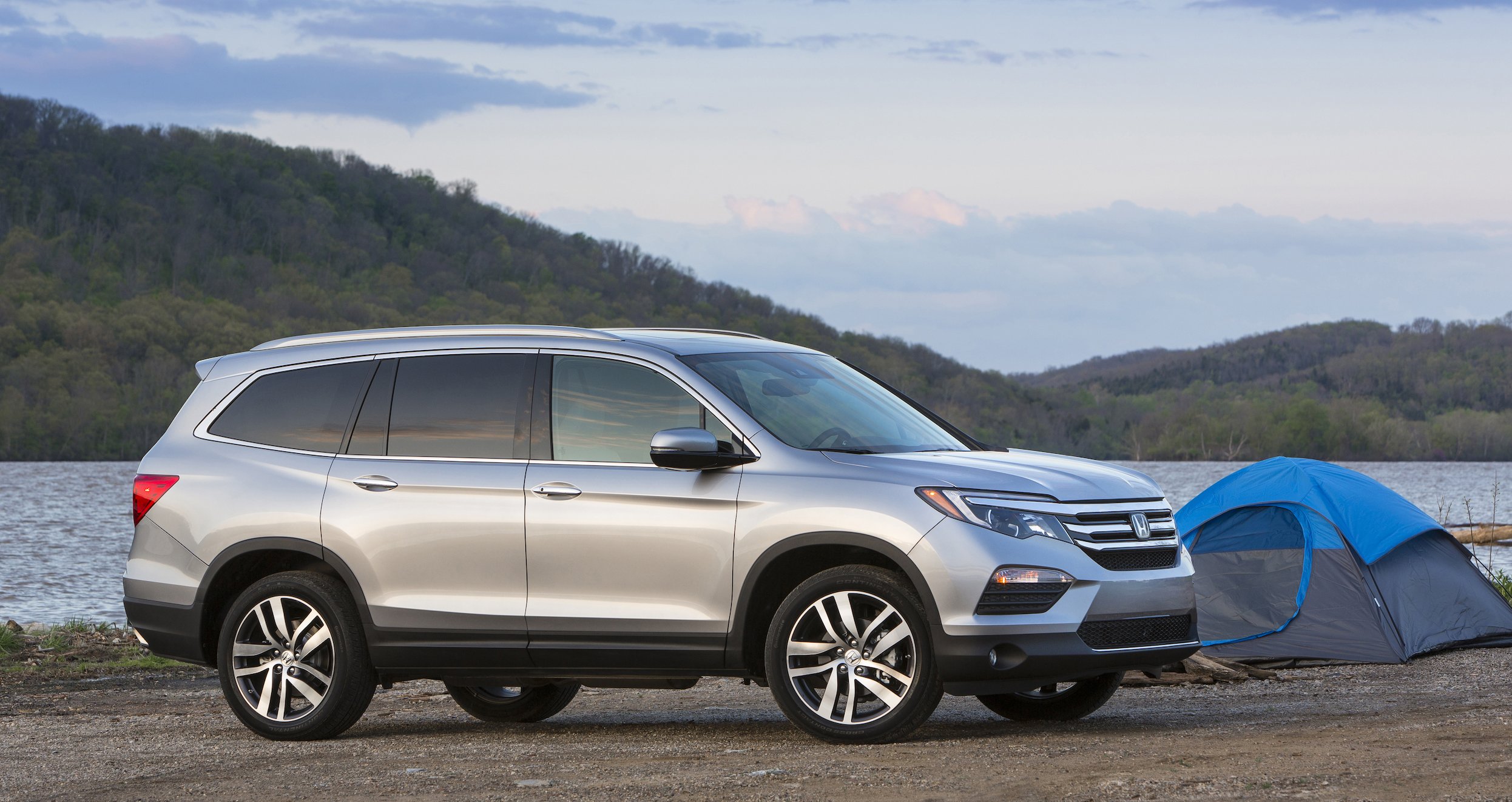 A silver 2018 Honda Pilot, a midsize SUV, parked next to a lakeside camp site. 